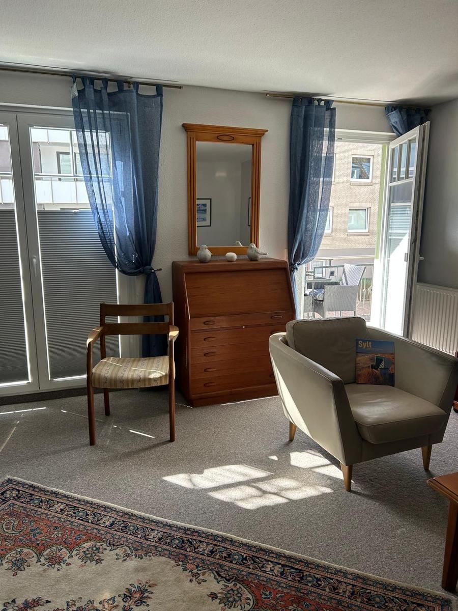 Ein Raum mit einem Holzschrank, einem Sessel und einem Stuhl. Vorhänge und Fenster sind sichtbar.