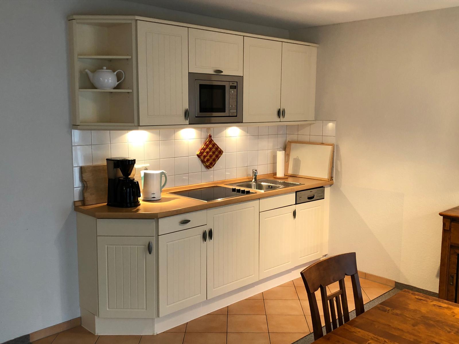 Küche mit weißen Schränken, Herd, Spüle und Mikrowelle. Holztisch und Stuhl sind sichtbar.
