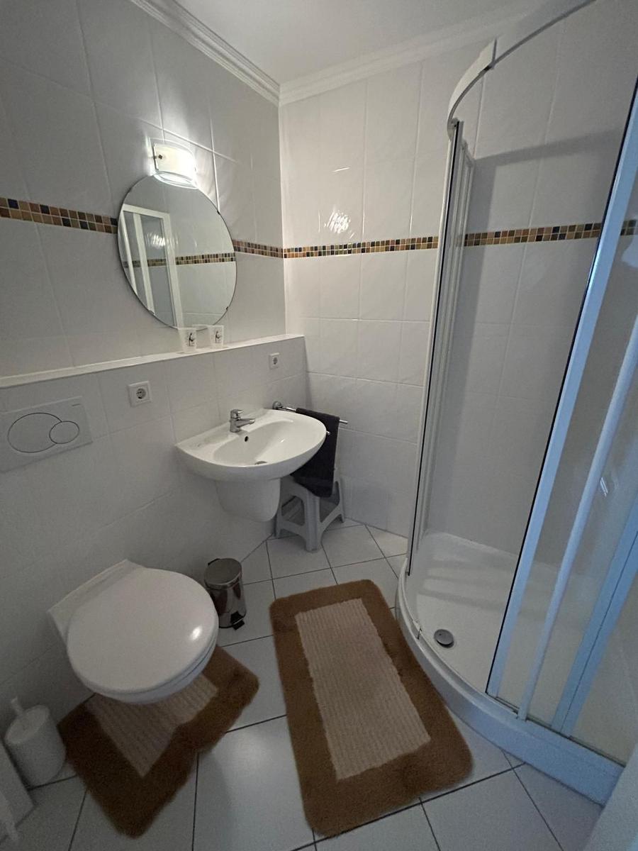 Badezimmer mit Dusche, Waschbecken und Toilette. Weiße Fliesen und runde Spiegel.