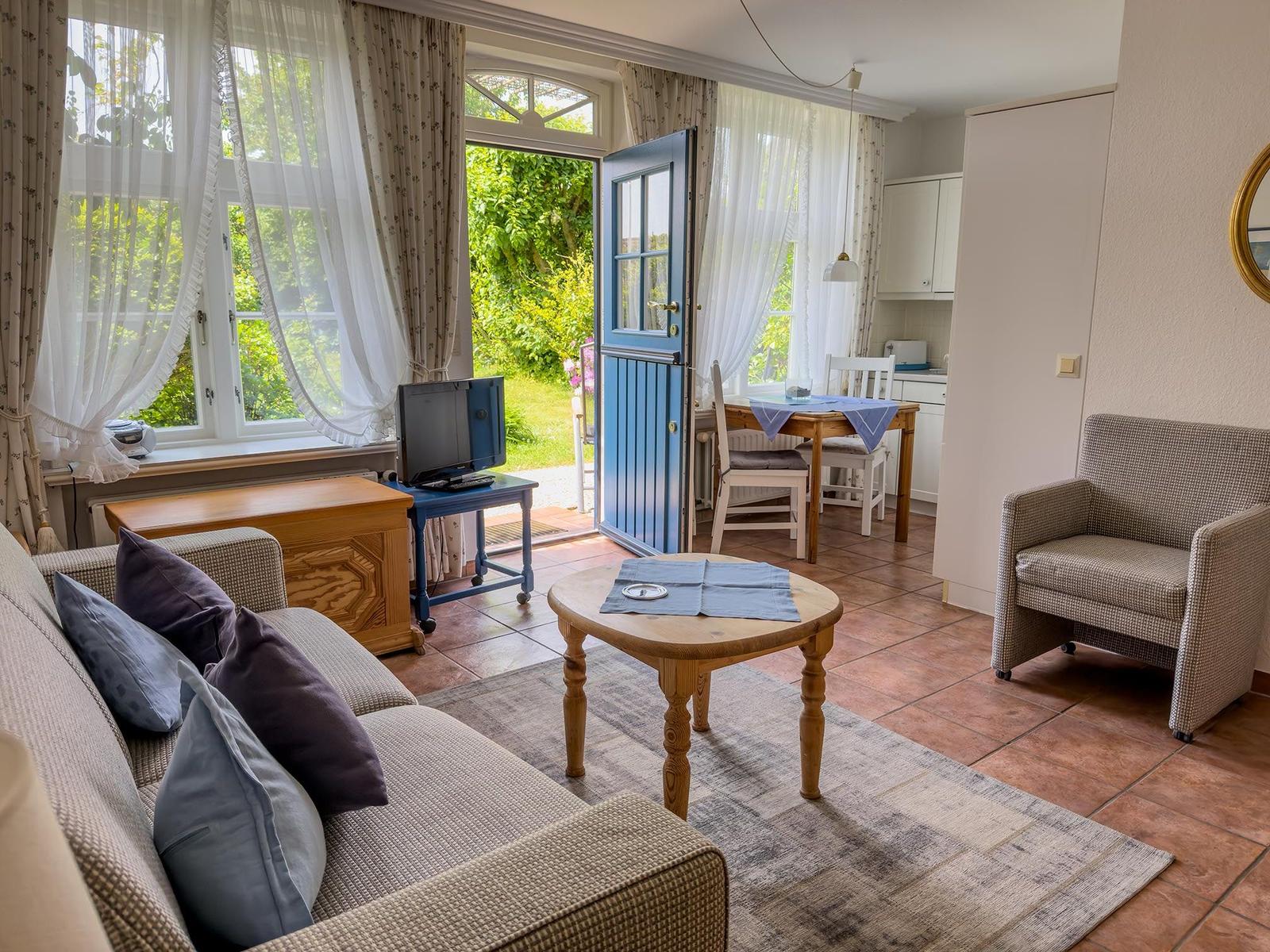 Wohnzimmer mit Sofa, Tisch und Stühlen. Durchgehendes Fenster und Tür zum Garten.