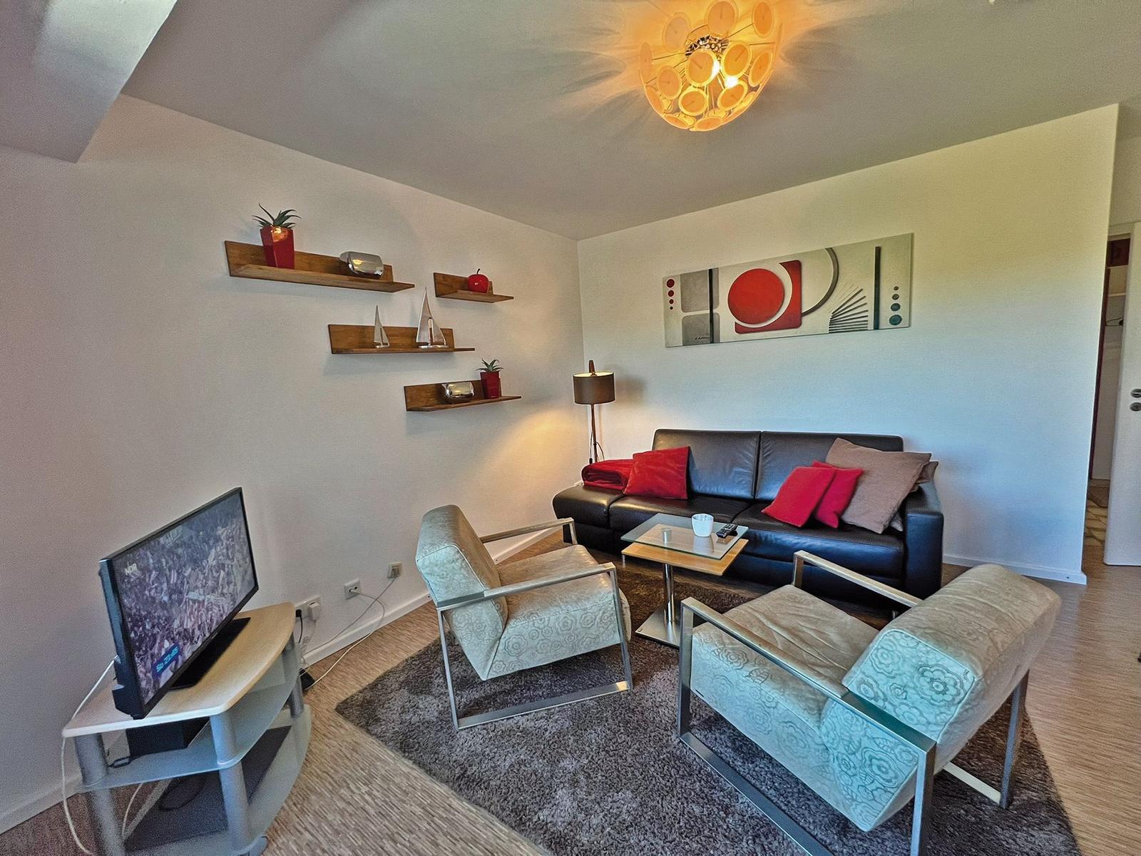 Wohnzimmer mit Sofa, zwei Sesseln, Fernseher und Wandregalen.