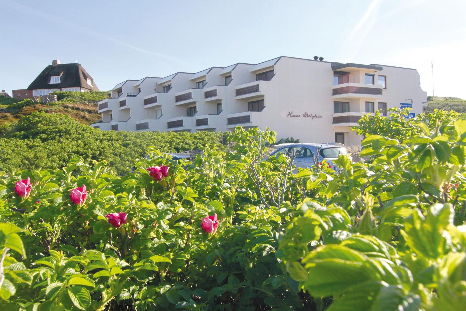Haus Dolphin mit Balkonen, umgeben von grüner Vegetation und roten Blüten.