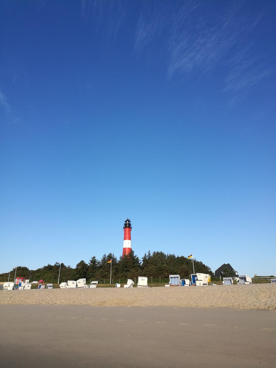 Roter Leuchtturm auf einer sandigen Küstenlandschaft mit Strandkörben und Bäumen im Hintergrund.