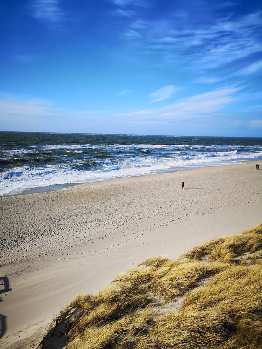 Weiterer Blick auf den Strand mit Wellen und Sanddünen im Vordergrund.