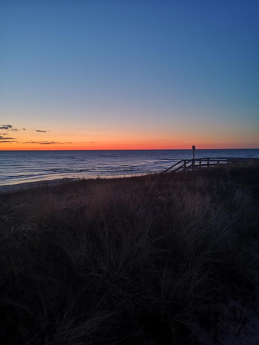 Sonnenuntergang über dem Meer mit Dünen und Holzsteg im Vordergrund.