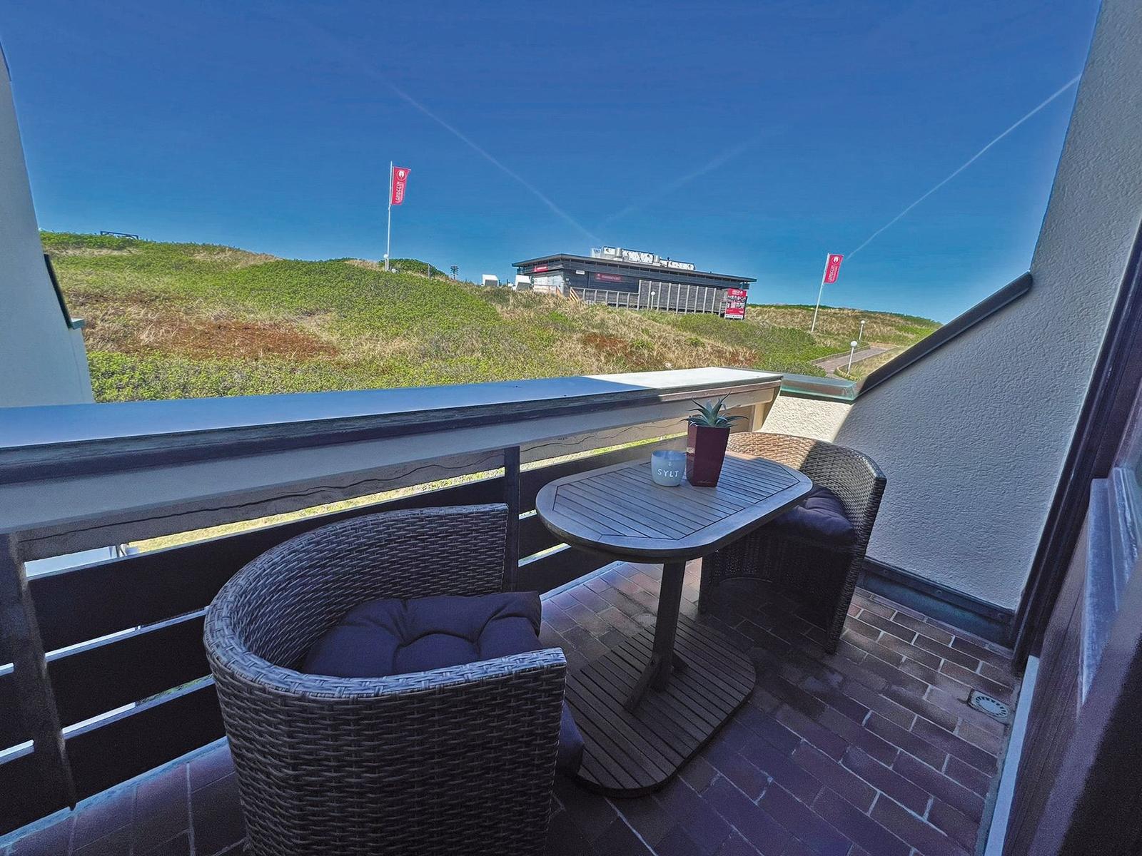 Terrasse mit Tisch und zwei Stühlen. Blick auf grüne Hügel und Gebäude.