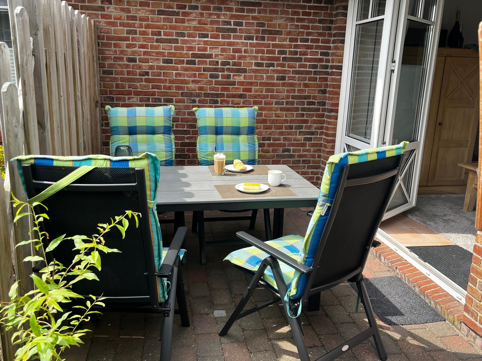 Terrasse mit Tisch und vier Stühlen, blau-grüne Kissen, Backsteinwand im Hintergrund.