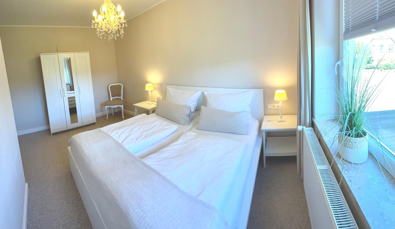 Schlafzimmer mit Doppelbett, Nachttischen, Kleiderschrank und Fenster mit Pflanze.