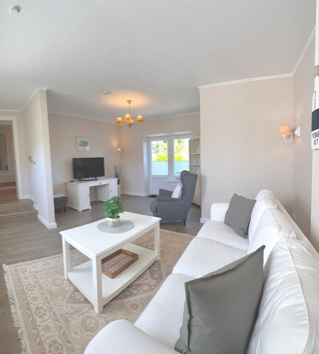 Wohnzimmer mit weißem Sofa, Couchtisch und Fernseher. Durchgang zur Küche und Schlafzimmer.
