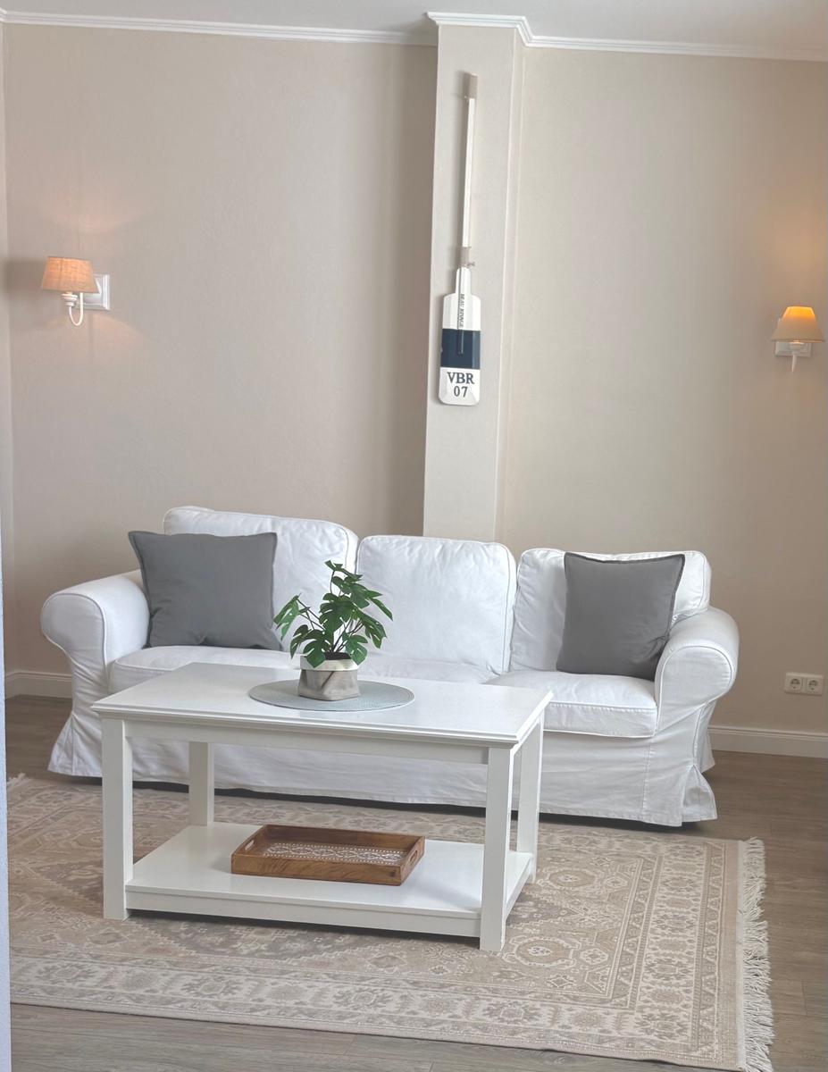 Ein Wohnzimmer mit weißem Sofa, Tisch und Teppich. An der Wand hängt ein Ruderboot.