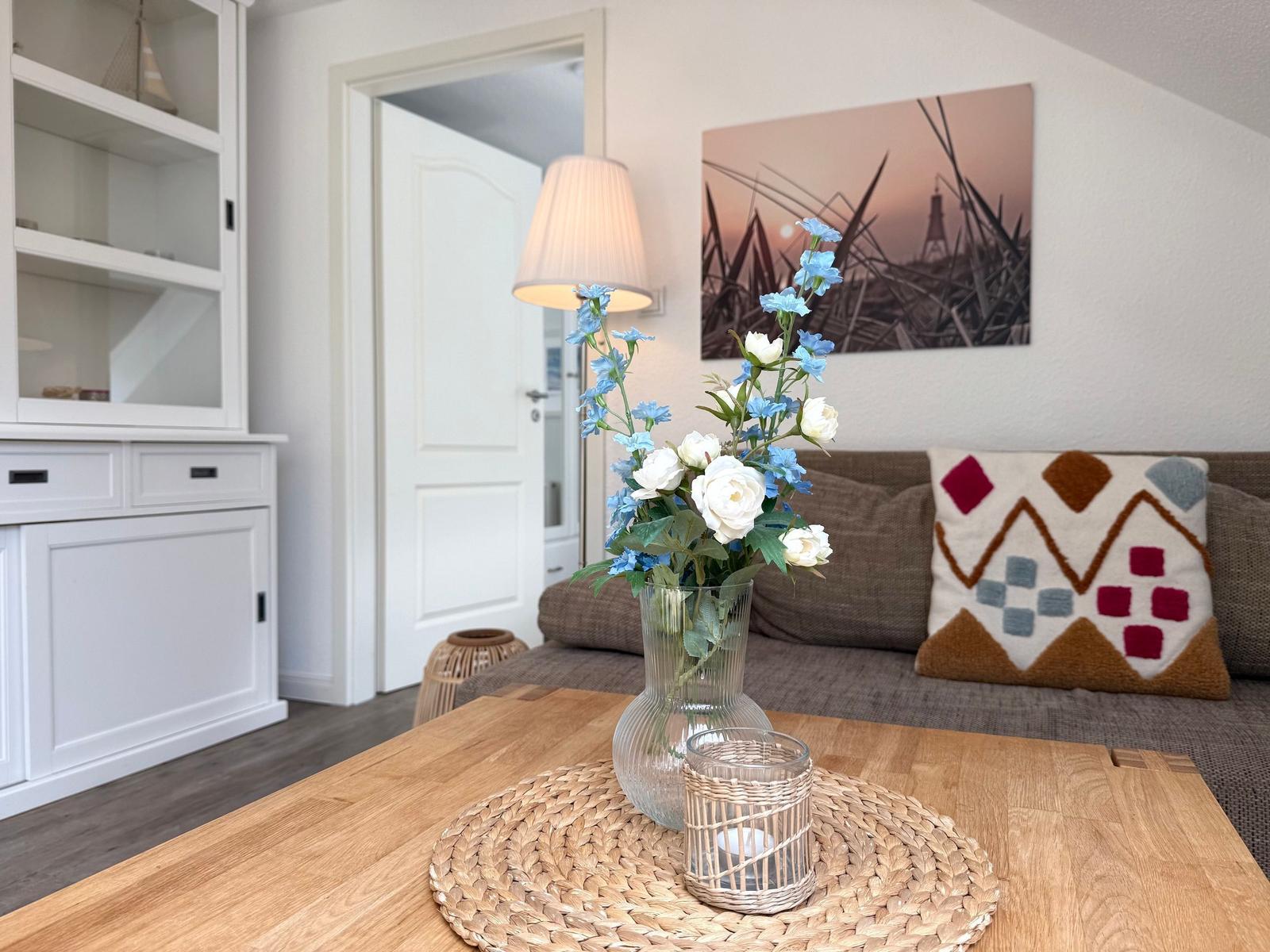 Ein Wohnzimmer mit Sofa, Tisch und Blumenarrangement. Wandbild und weiße Einbauschränke sind sichtbar.