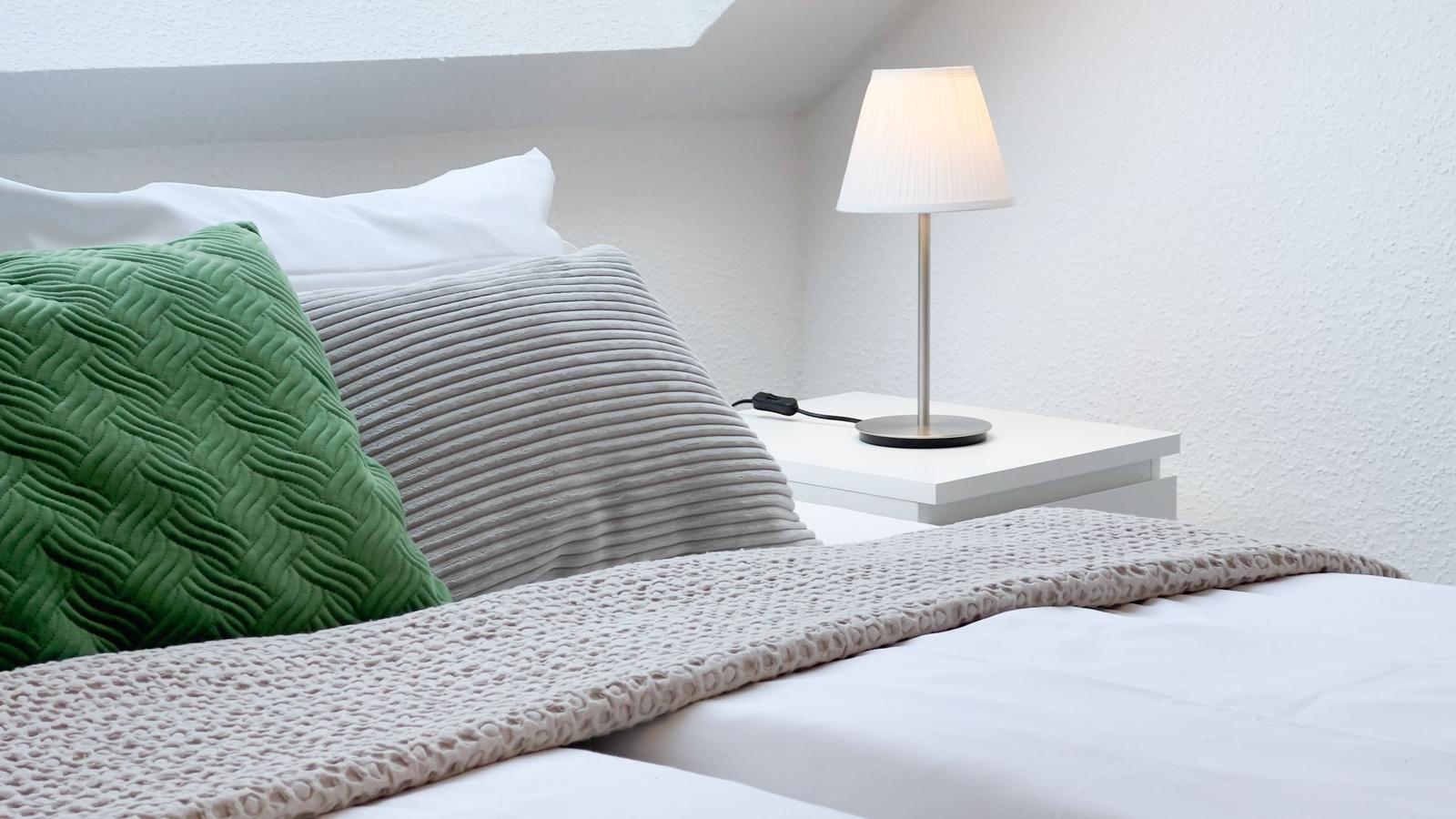 Doppelbett mit grünem und grauem Kissen, weißem Nachttisch und Lampe.