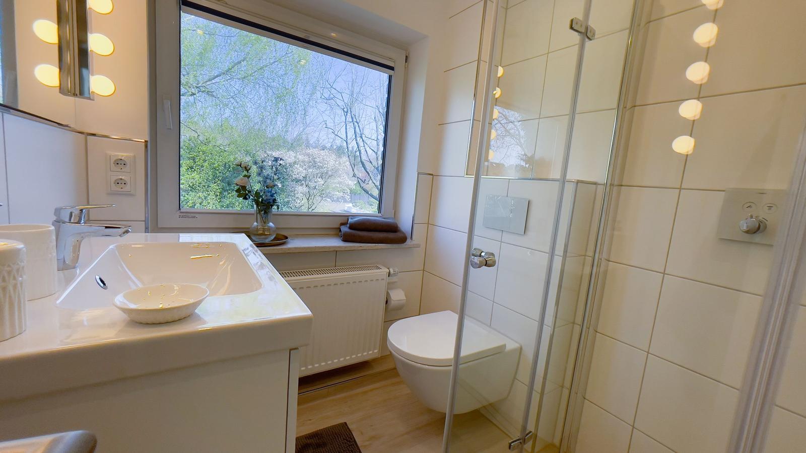 Badezimmer mit Dusche, Waschbecken und Fensterblick auf Grün.