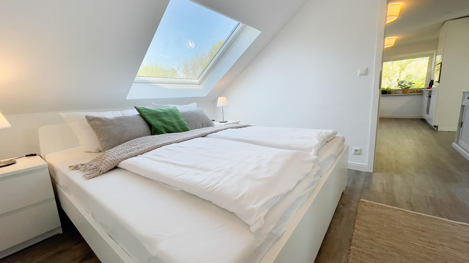 Schlafzimmer mit Dachschräge, großem Fenster und weißem Bett mit Kissen.