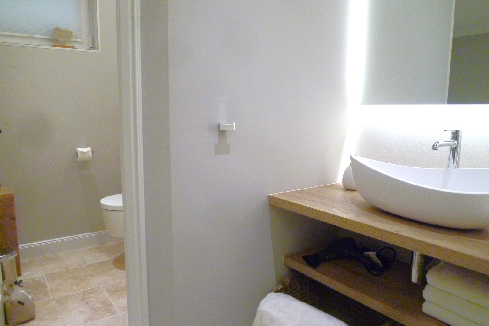 Bad mit Waschbecken auf Holzunterschrank, Spiegel und Toilette im Hintergrund.