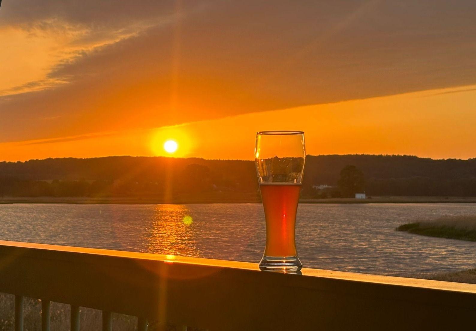 Ein Bierglas steht auf einer Terrasse vor einem See bei Sonnenuntergang.