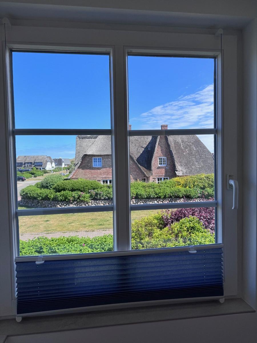 Durch das Fenster blickt man auf ein traditionelles Haus mit Strohdach und grüne Beete.
