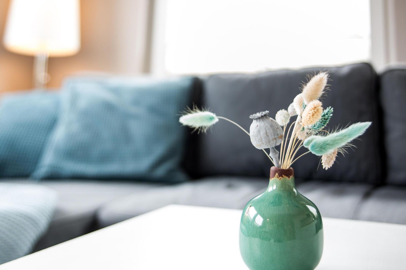 Ein grüner Vasen mit Trockenblumen steht auf einem Tisch vor einem Sofa.