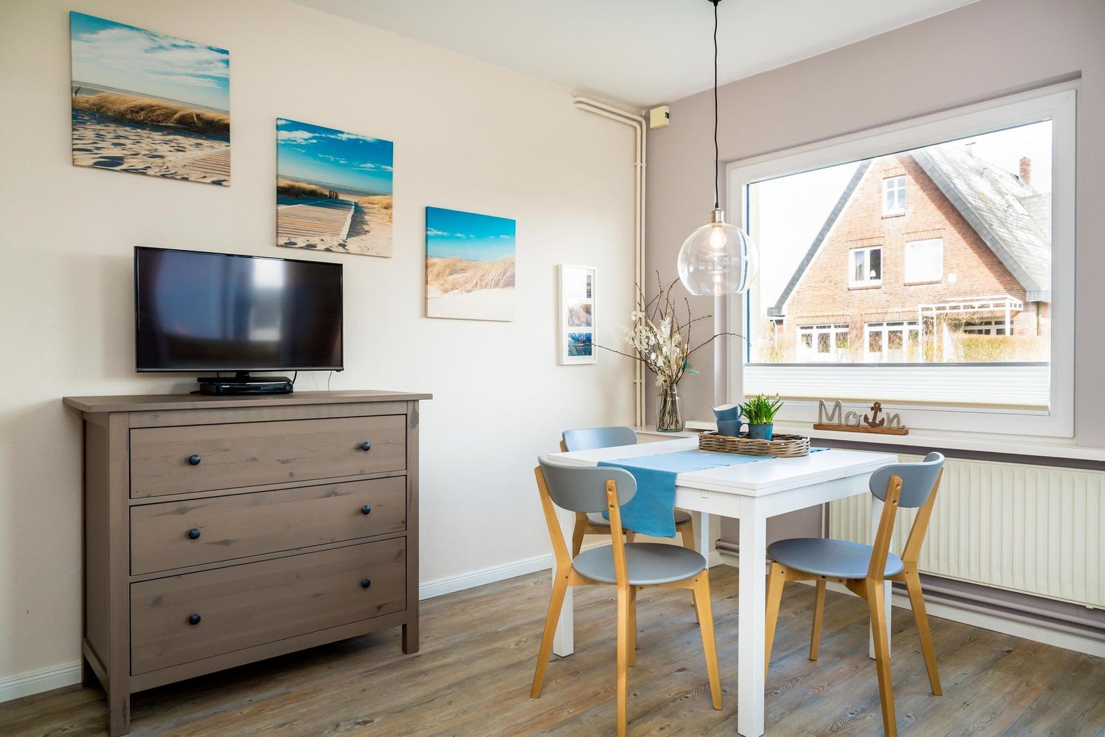 Essbereich mit Tisch, Stühlen und Fernseher. Fensterblick auf ein Haus.