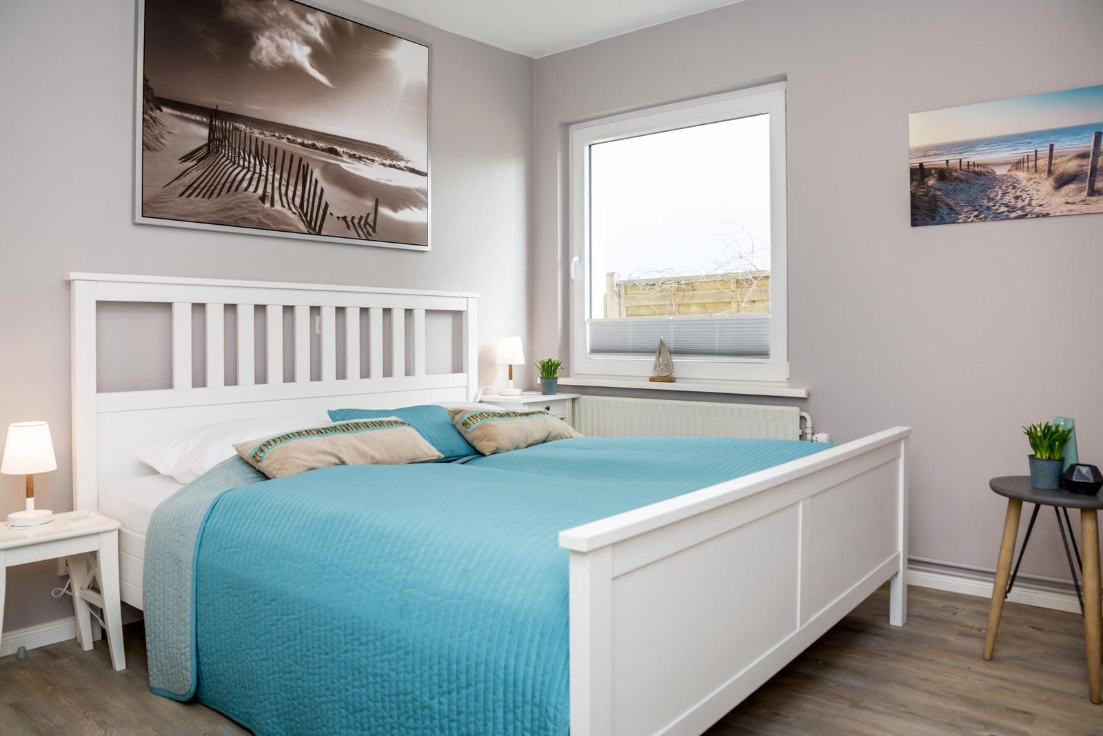 Schlafzimmer mit weißem Bett, blauer Bettdecke und Fenster mit Blick auf Strand.