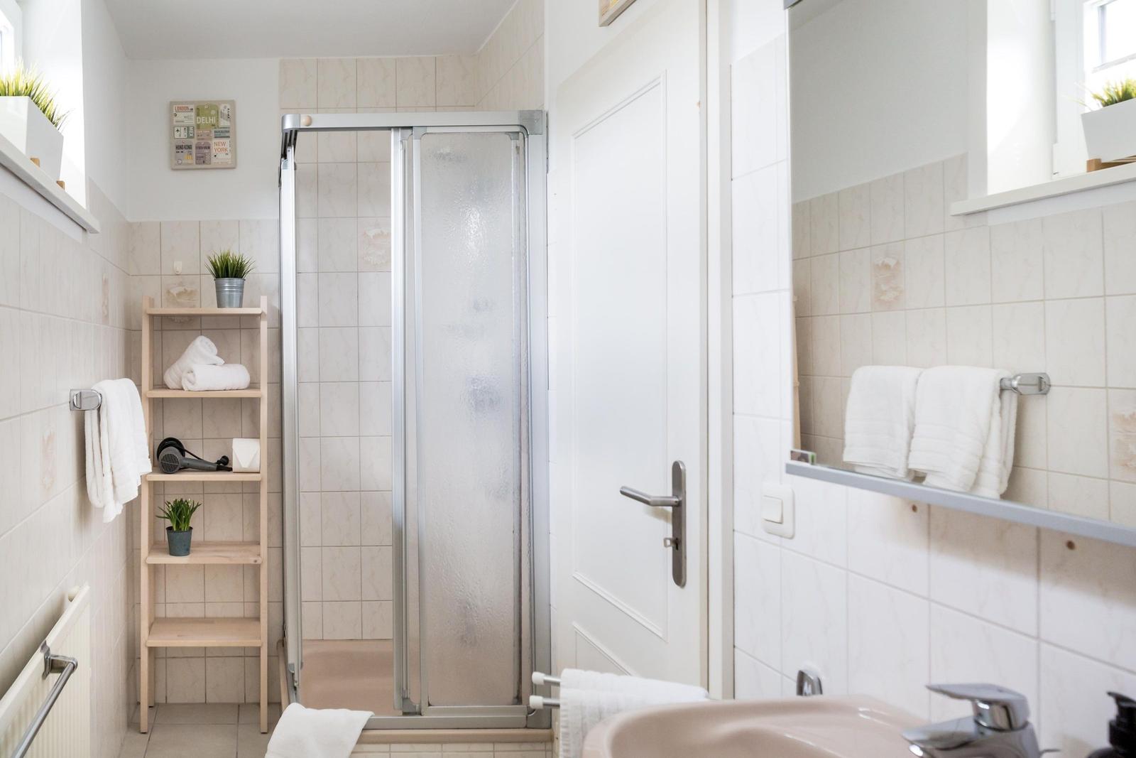Badezimmer mit Dusche, Waschbecken und Holzregal mit Handtüchern und Pflanzen.