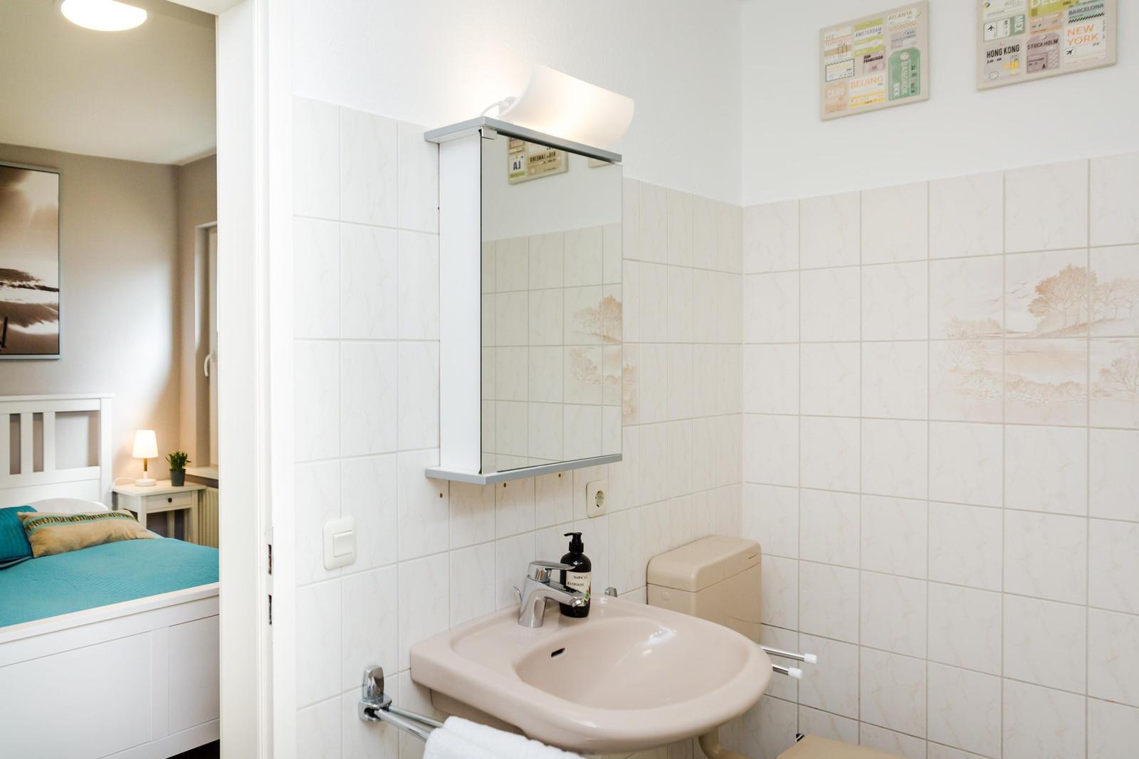 Badezimmer mit Waschbecken, Spiegel und Toilette. Blick in das Schlafzimmer mit Bett.