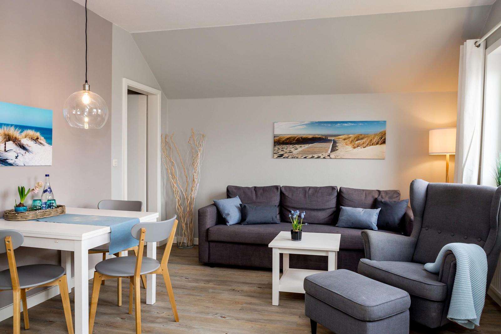 Wohn- und Essbereich mit Sofa, Stuhl und Tisch. Wandbilder zeigen Strandlandschaften.
