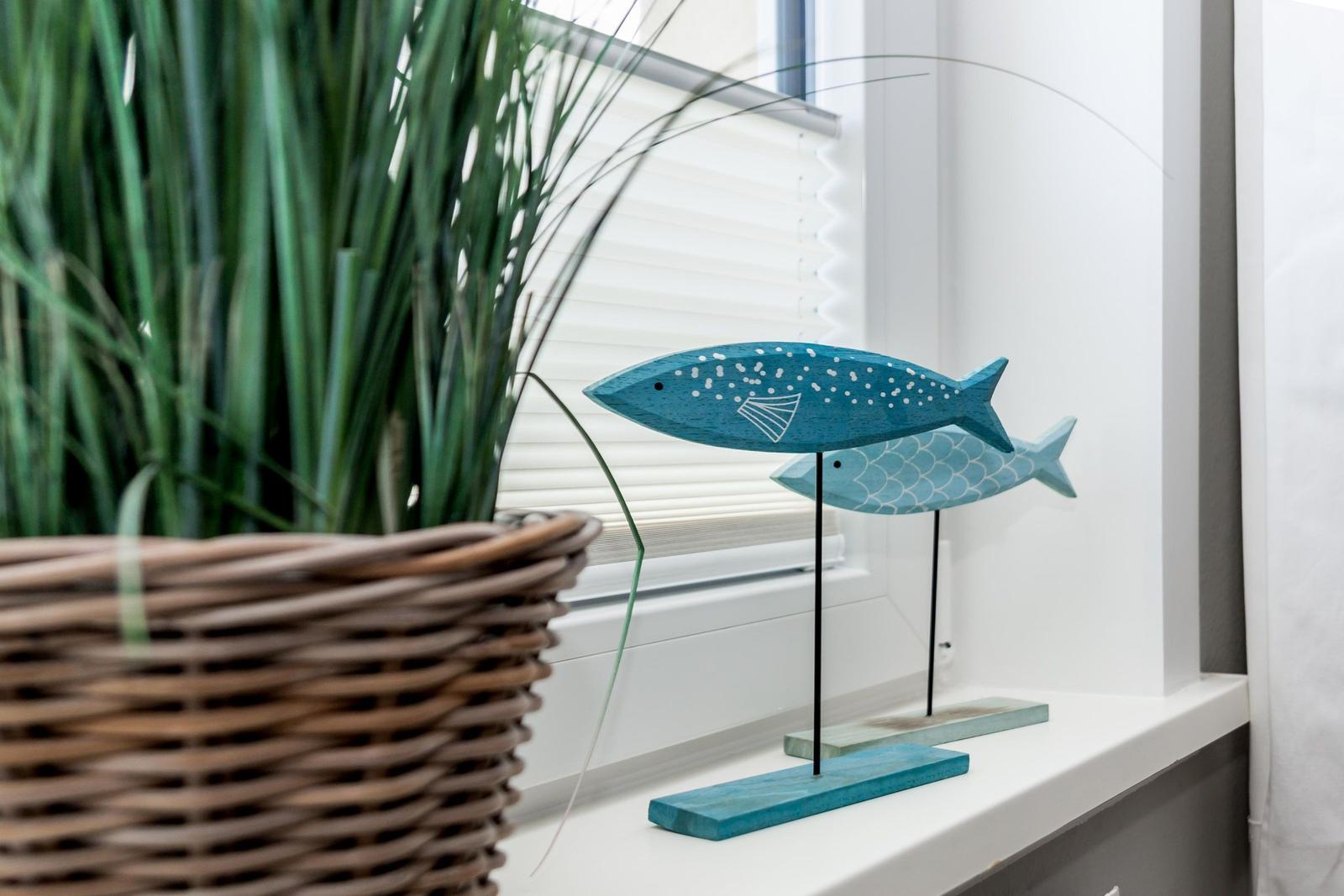 Zwei hölzerne Fische auf einem Fensterbrett neben einer Pflanze in einem Korb.