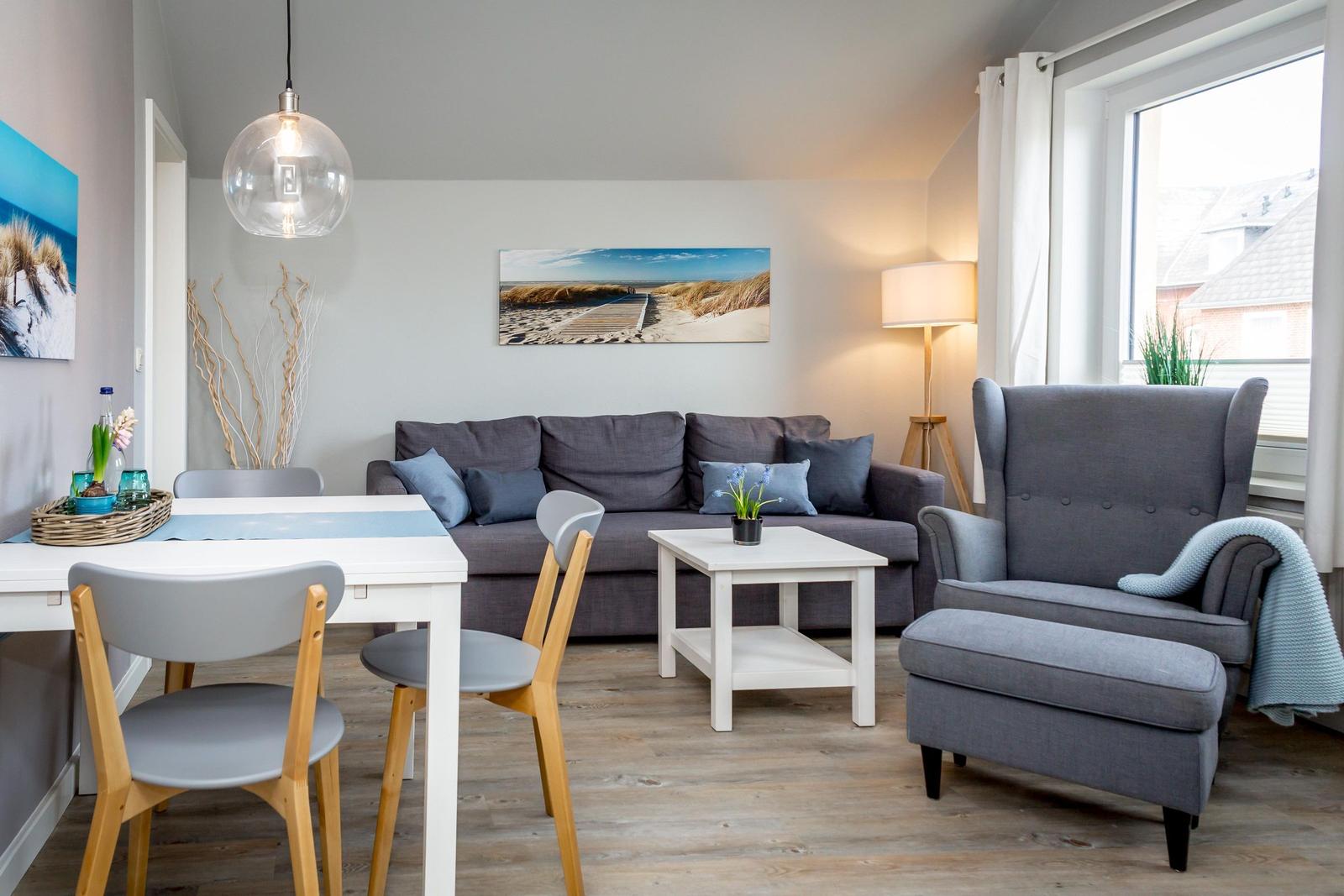 Wohn- und Essbereich mit grauem Sofa, Esstisch und Sessel. Holzboden und große Fenster.