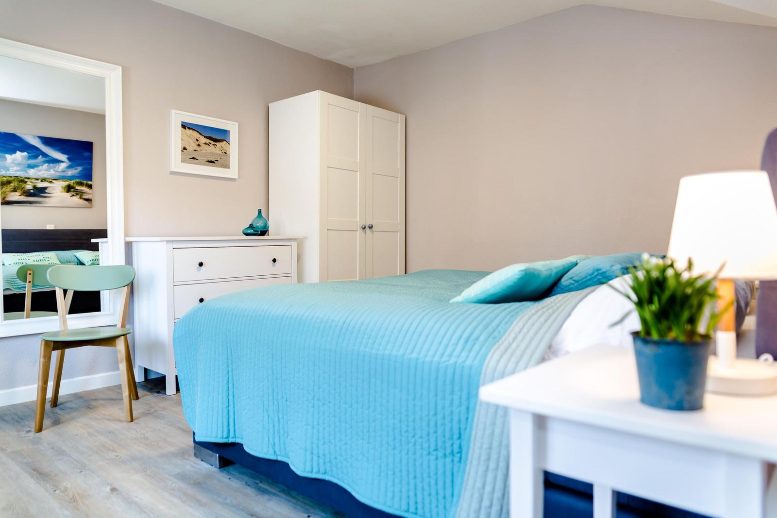 Schlafzimmer mit Bett, Schrank und Nachttisch. Wandbild und Pflanze sichtbar.