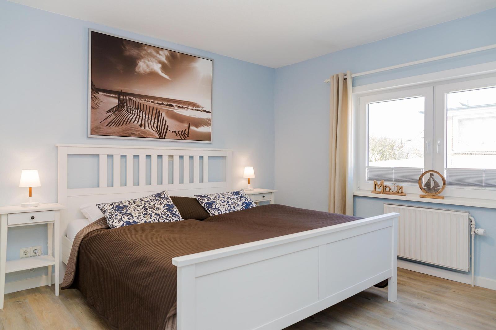 Schlafzimmer mit weißem Bett, Wandbild und Fenster mit Vorhängen.