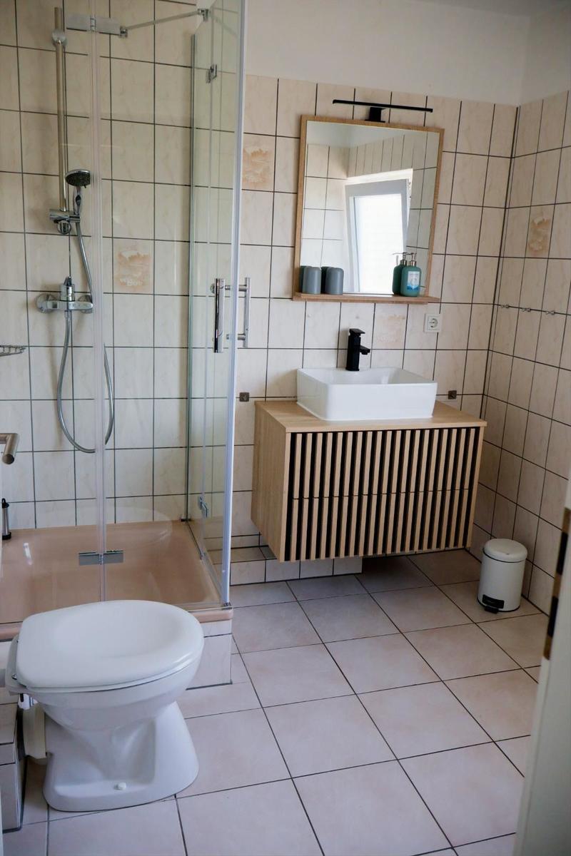 Badezimmer mit Dusche, Waschbecken und Toilette. Holzunterschrank und Spiegel.