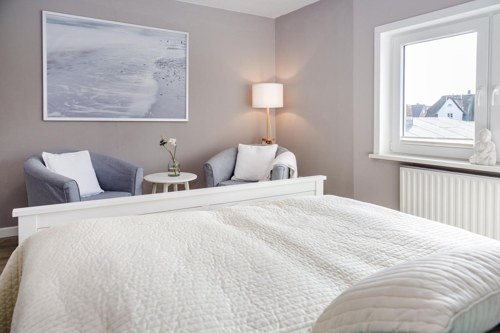 Schlafzimmer mit Bett, zwei Sesseln, Tisch und Fenster. Wandbild und Stehleuchte.