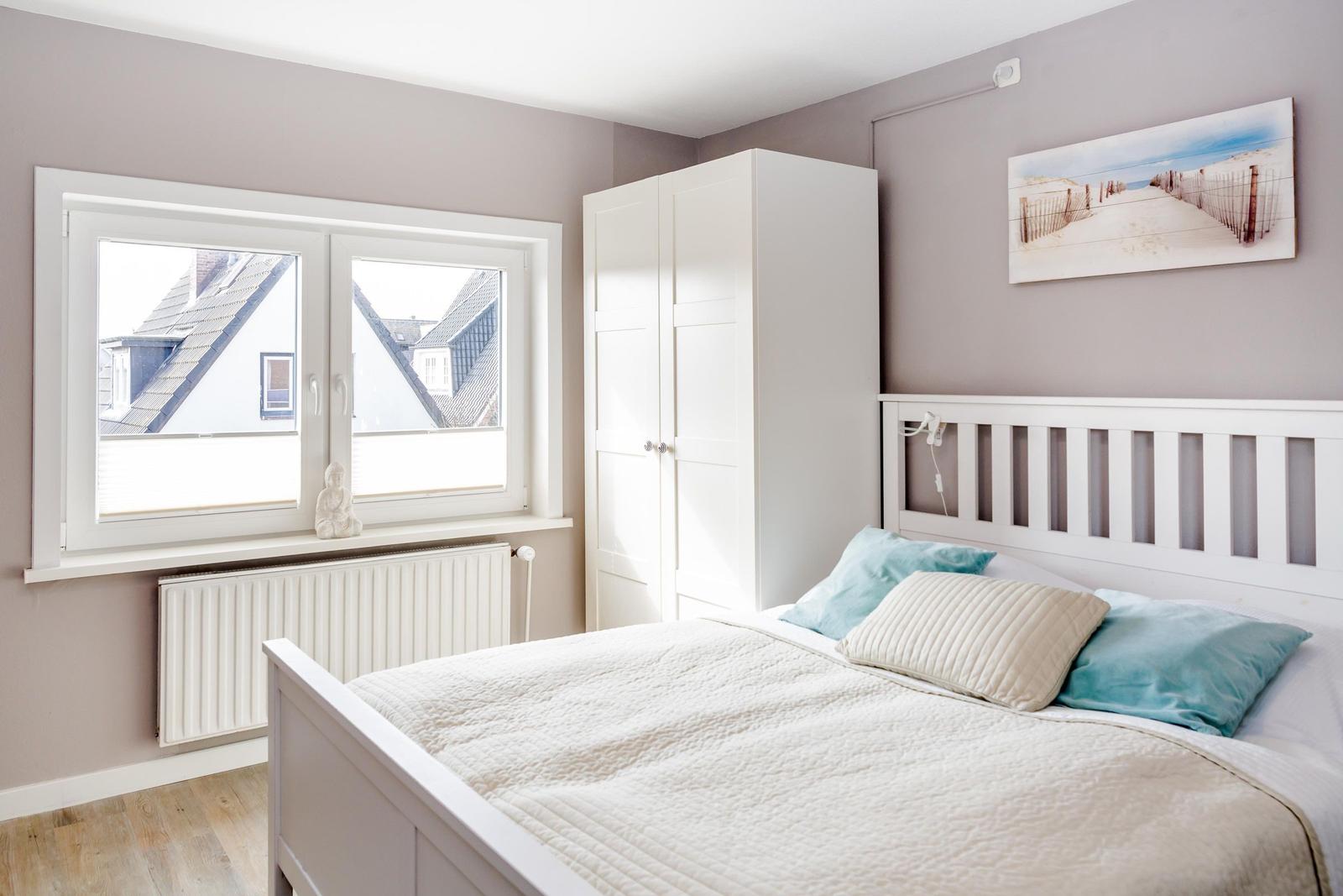 Schlafzimmer mit weißem Bett, Kleiderschrank und Fensterblick auf Dächer.