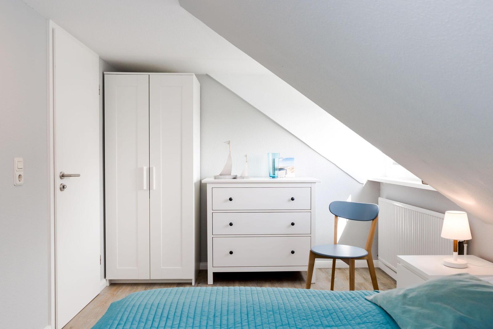 Bedroom with sloped ceiling, white dresser, chair, and bed with blue bedding.