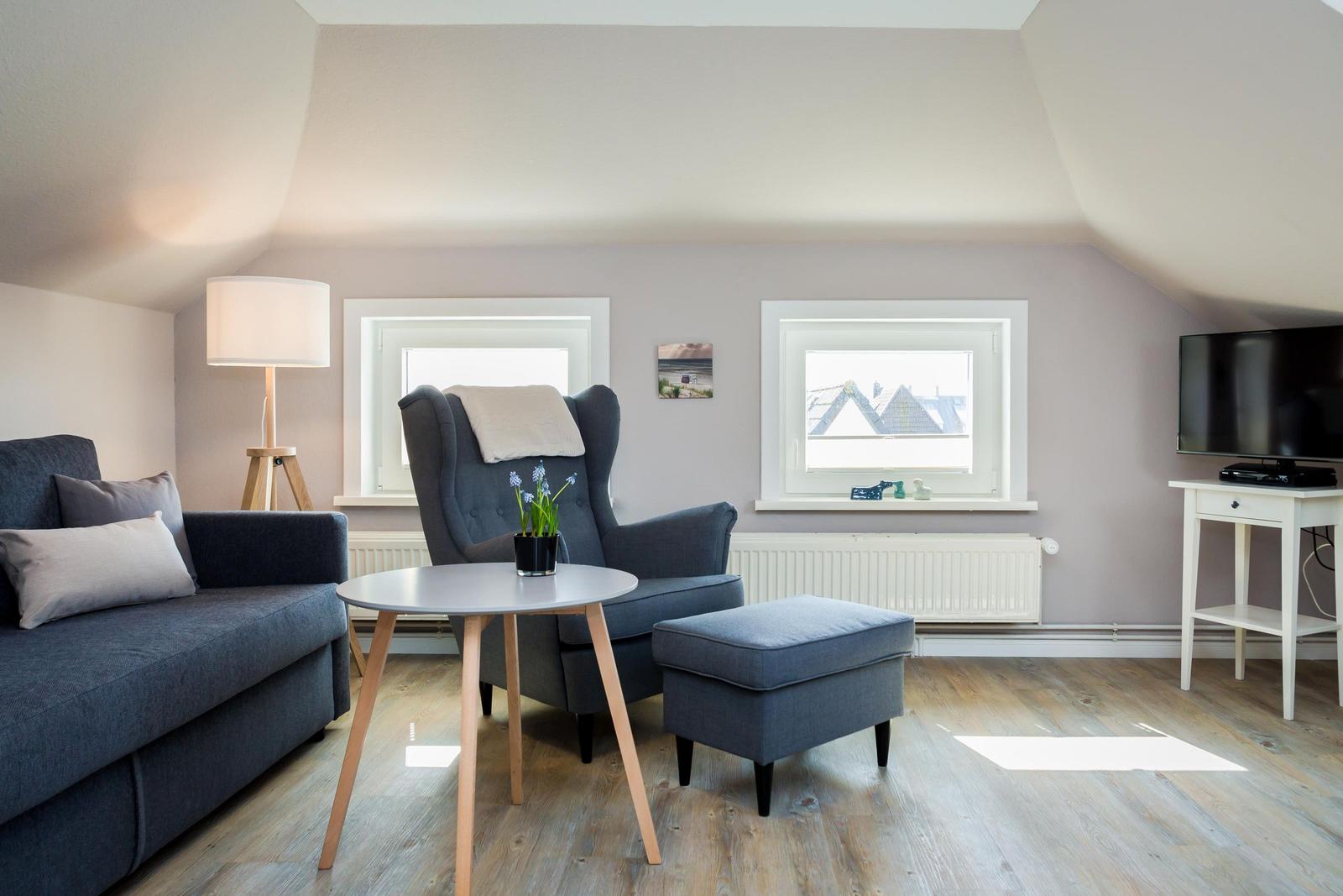 Living room with sofa, armchair, table, and TV. Wooden floor and two windows.