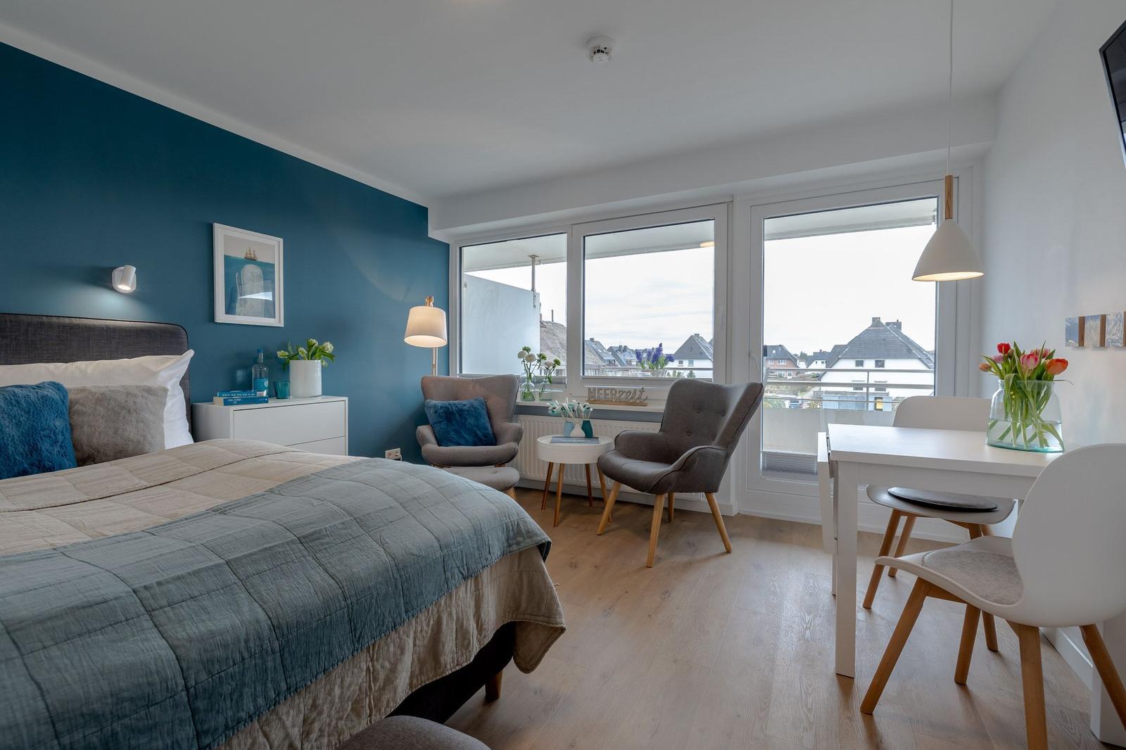 Schlafzimmer mit Bett, Sitzgelegenheiten und Tisch. Blick auf Balkon und Dachlandschaft.