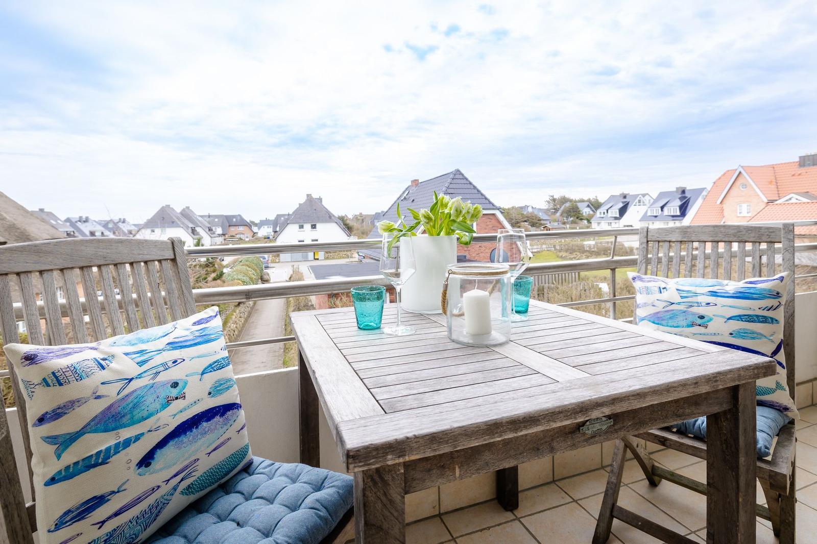 Terrasse mit Holztisch, Stühlen und Fischmotiv-Kissen. Blick auf Wohnhäuser und Himmel.