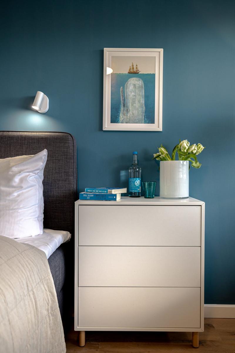 Schlafzimmer mit Nachttisch, Bett und Wandbild. Auf dem Tisch stehen Bücher, eine Flasche und Blumen.