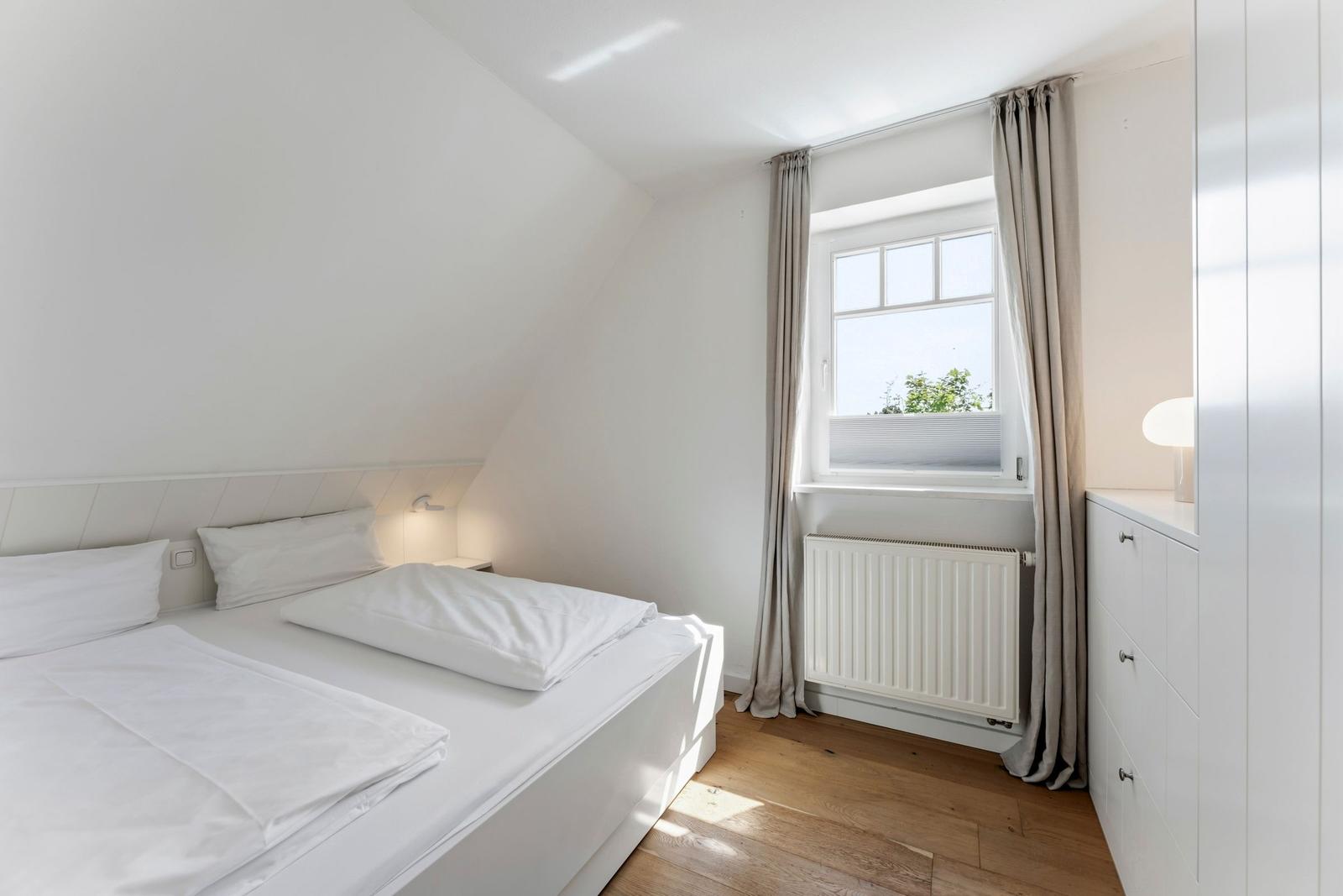 Schlafzimmer mit Dachschräge, weißem Bett, Fenster mit Vorhängen und Holzboden.