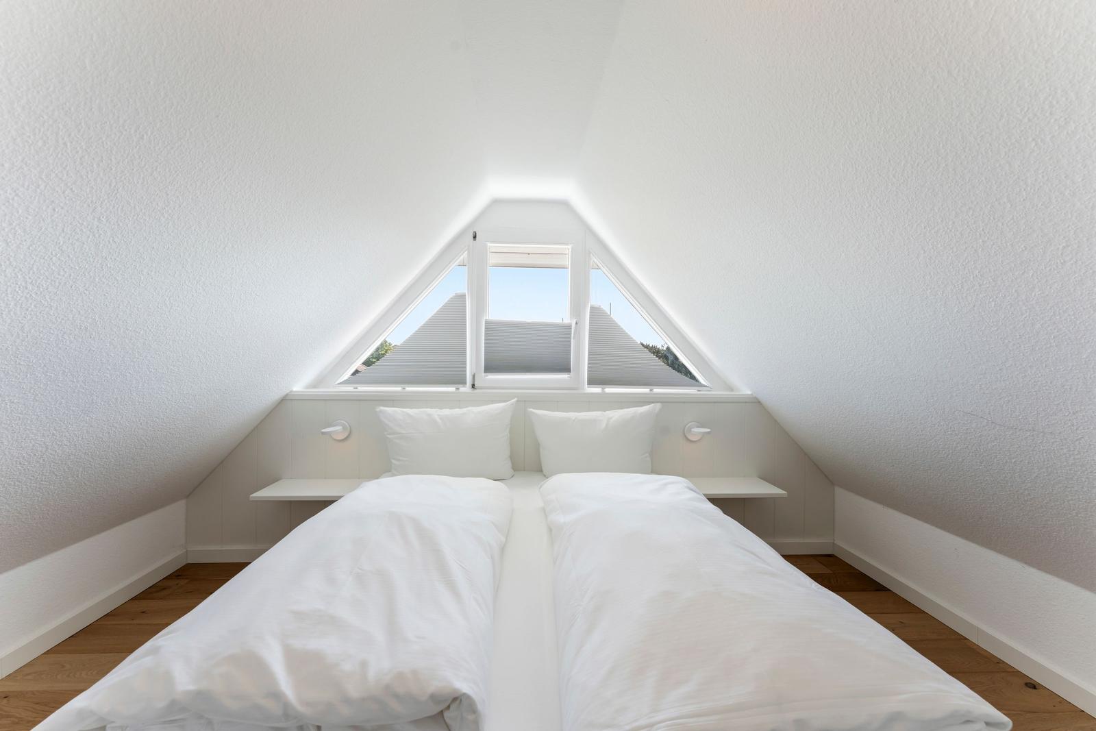 Zimmer mit Doppelbett unter Dachschräge und Dreifachfenster mit Blick auf Dächer.