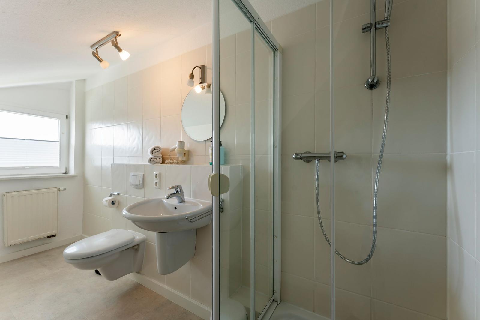 Badezimmer mit Dusche, Waschbecken und Toilette. Fenster und Heizkörper sichtbar.
