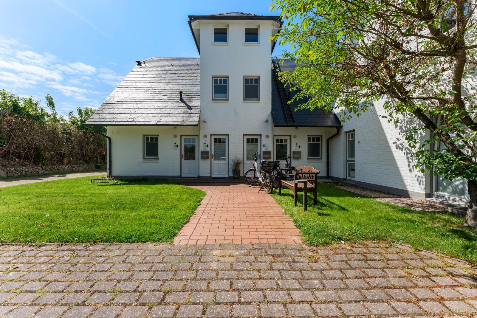 Weißes Haus mit Garten, Terrasse und Fahrrädern. Pflasterweg führt zur Eingangstür.
