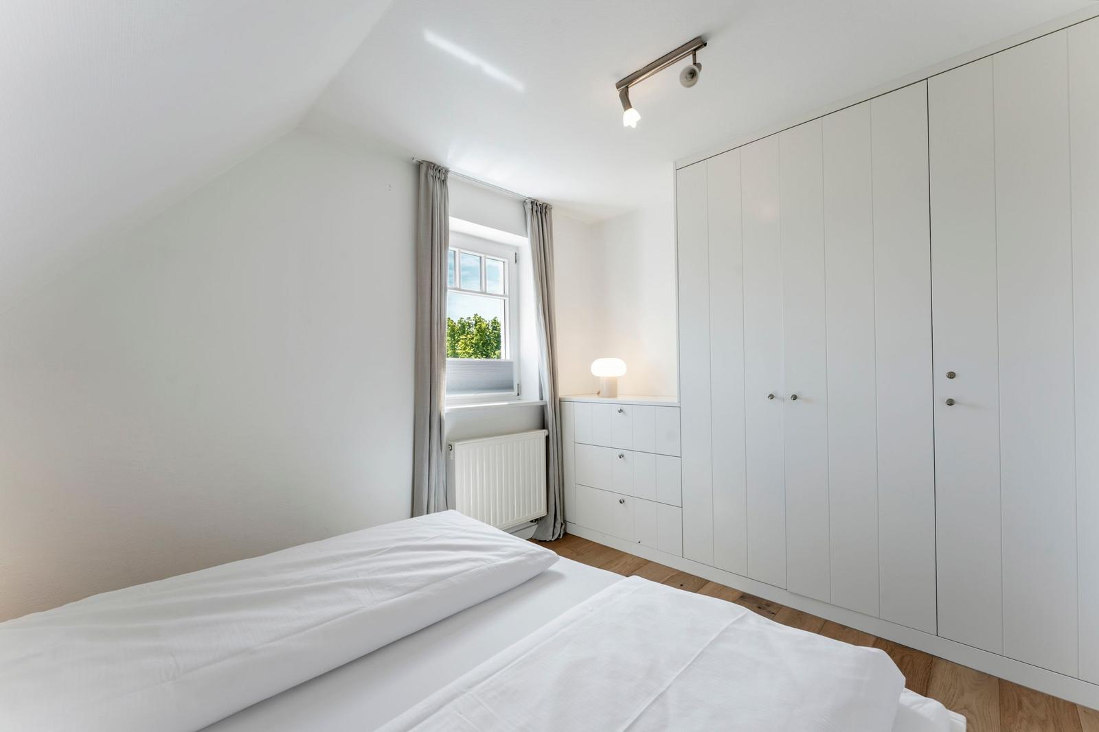 Schlafzimmer mit Dachschrägen, weißem Bett, Kleiderschrank und Fenster mit Blick auf Bäume.