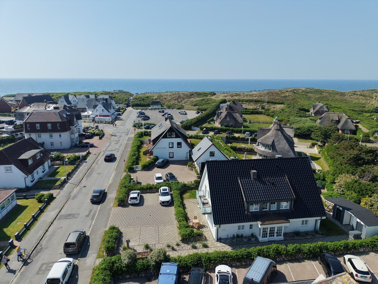 Haus mit dunklem Dach, umgeben von Grünflächen und Parkplätzen. Im Hintergrund liegt das Meer.