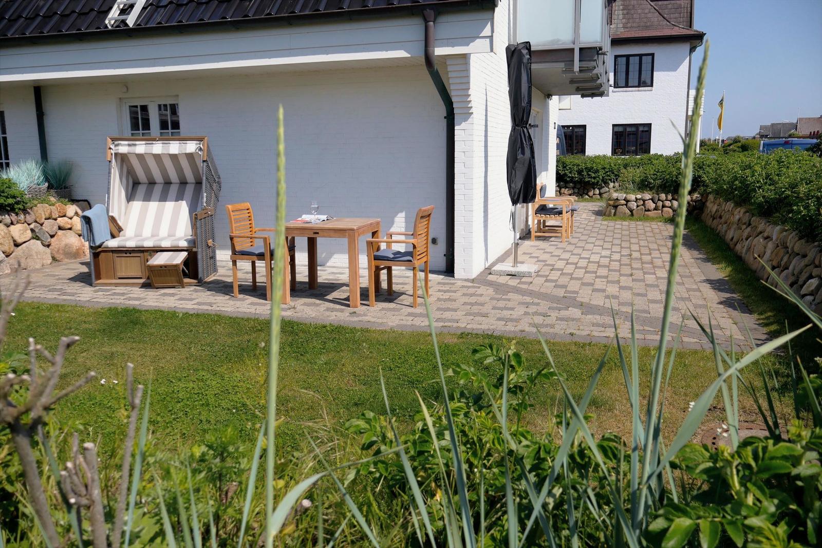 Terrasse mit Holztisch, Stühlen und Strandkorb. Hinterhof mit Steinmauer und Gras.