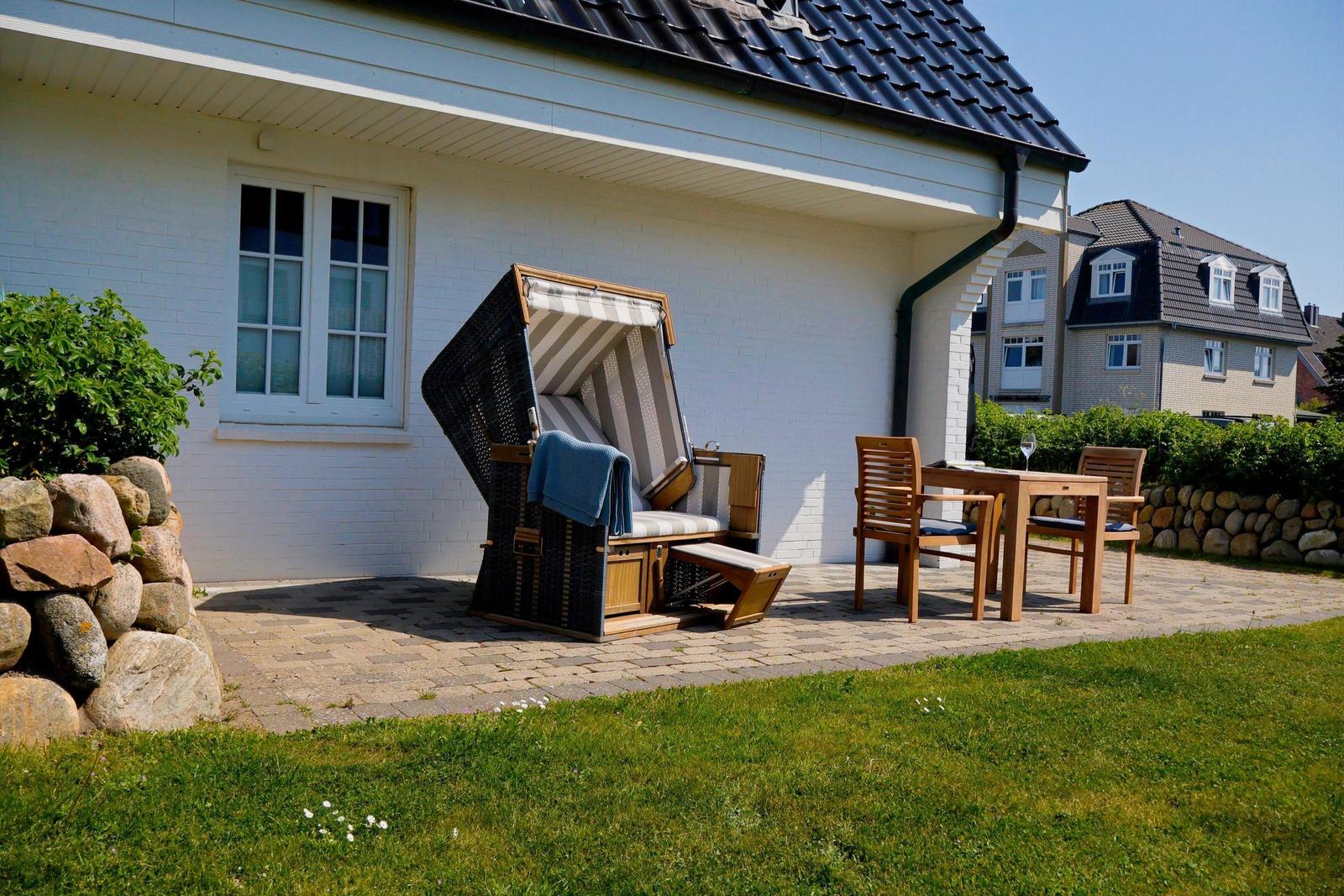 Terrasse mit Strandkorb, Tisch und Stühlen vor weißem Haus mit Steinmauer.
