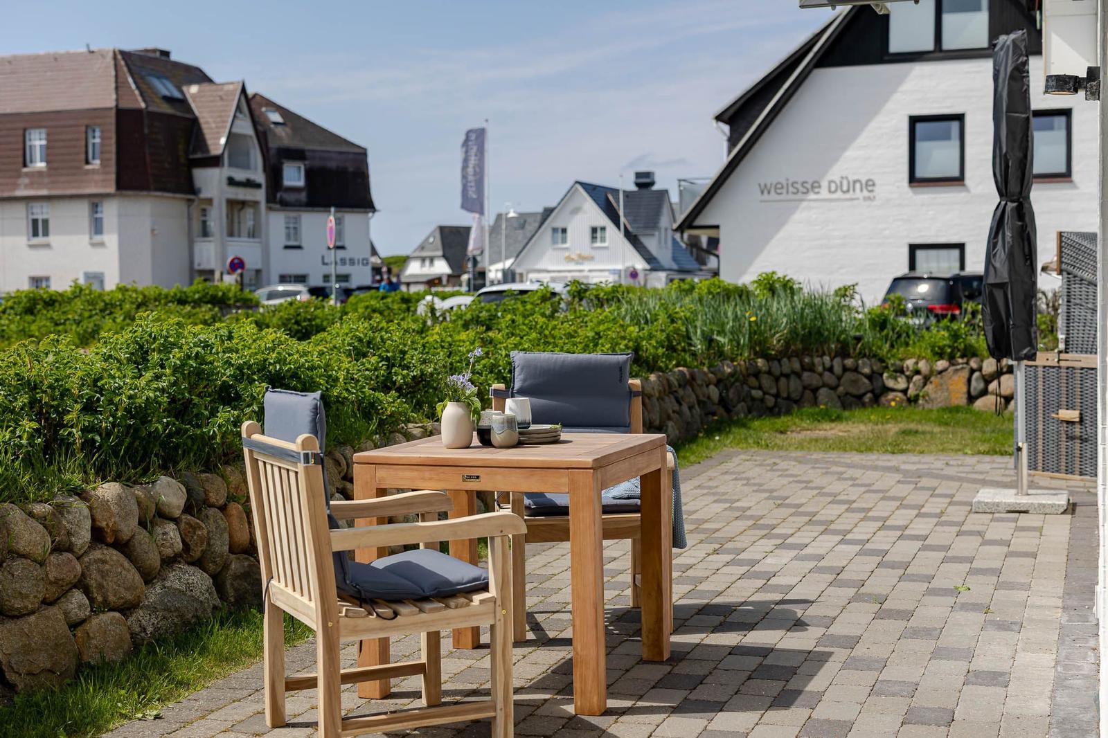 Terrasse mit Holztisch und Stühlen, Blick auf weiße Gebäude und Grünflächen.