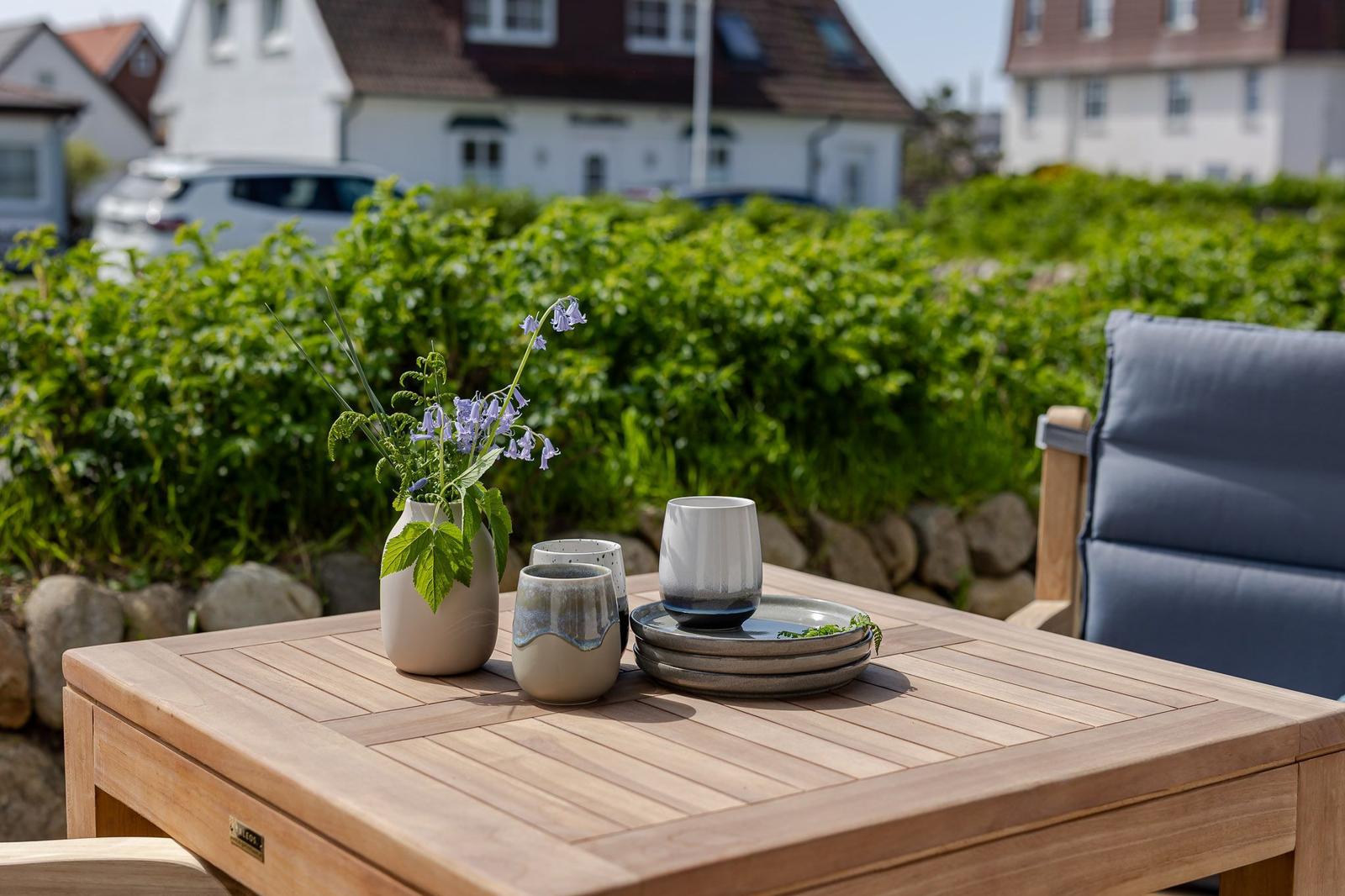 Holztisch mit Tassen, Tellern und Blumen im Garten. Hintergrund: grüne Hecke und Häuser.