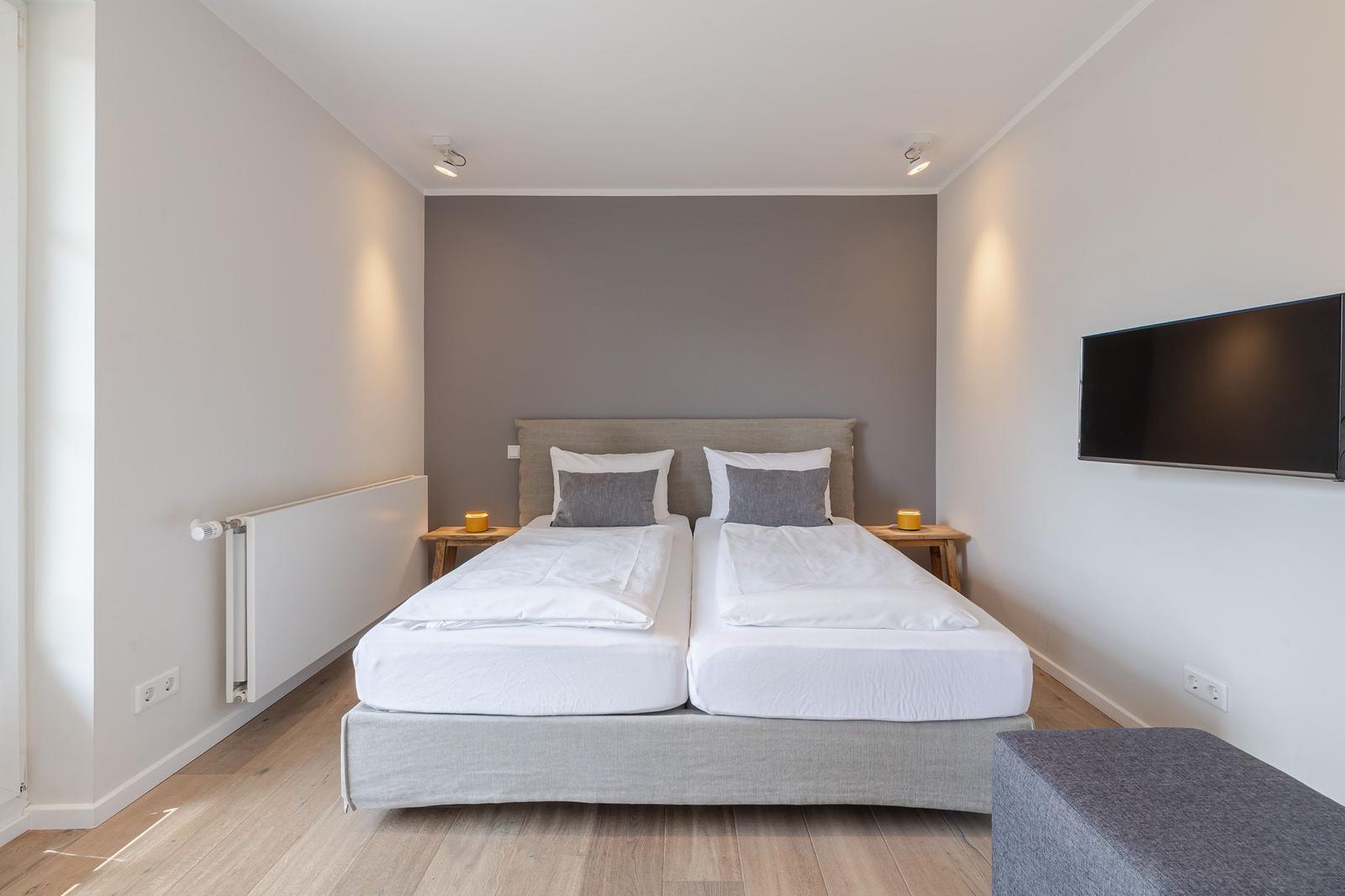 Doppelbett mit weißen Laken, zwei Nachttische und Wandfernseher in einem Schlafzimmer.